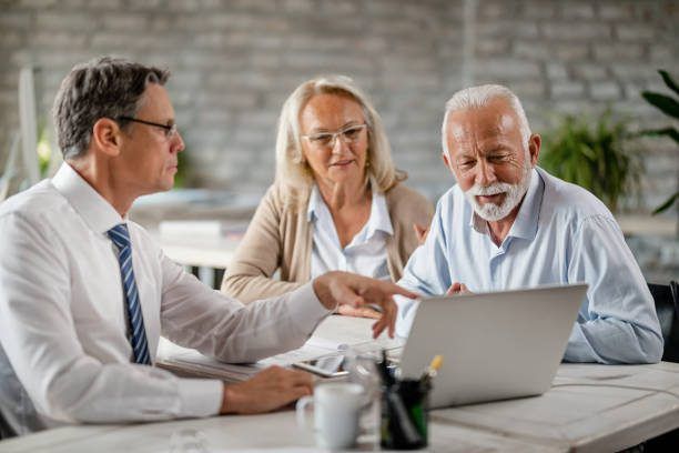 Senior couple using laptop with their financial advisor during a meeting int he office. Focus is on senior man.